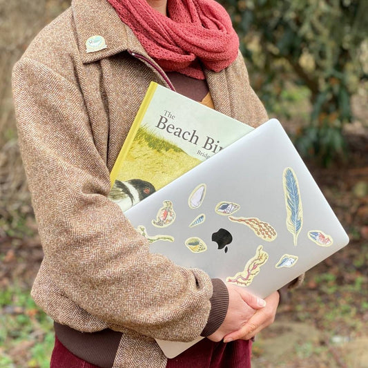 Person holding a laptop with nature-themed stickers and a book titled 'The Beach Birds' in an outdoor setting.