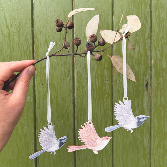 Three superb fairywren hanging ornaments  suspended from a eucalyptus branch held by a hand, set against a green wooden plank background.