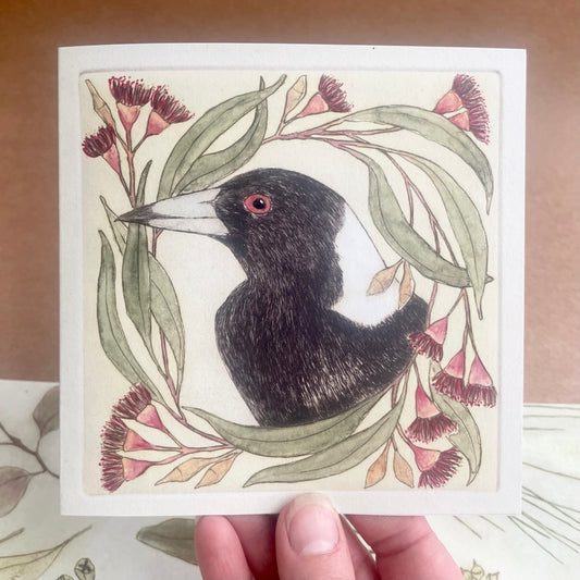 A Christmas card featuring a coloured drawing of a magpie surrounded by red and green foliage, with a blank interior, held in a person's hand.