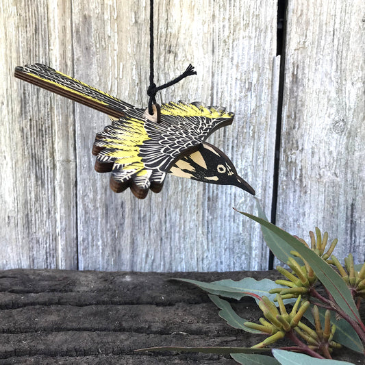 A wooden bird ornament, shaped like a black and yellow bird a new holland honeyeater, hanging from a black string against a weathered gray wooden fence background with eucalyptus leaves in the foreground.
