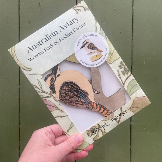 Hand holding a wooden bird figurine packaging with 'Australian Aviary' branding against a green wooden background.