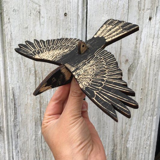 A hand holds a wooden bird cutout, an australian magpie with wings spread, featuring a distressed black finish and natural wood grain details, set against a weathered gray wood plank background.