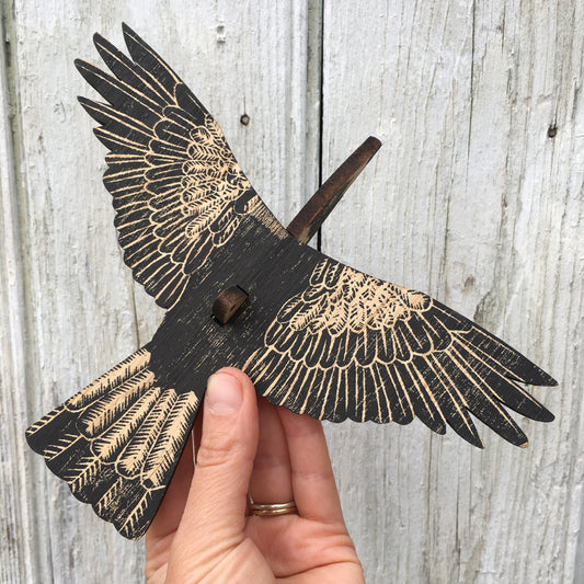 A hand holds a wooden bird cutout, an australian magpie with wings spread, featuring a distressed black finish and natural wood grain details, set against a weathered gray wood plank background.